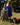 Woman in a blue dress holding a child, standing next to a brown dog on a leash in a park.