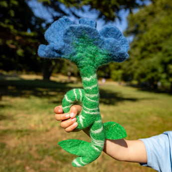 Hand holding a blue and green felt flower toy outdoors with trees in the background