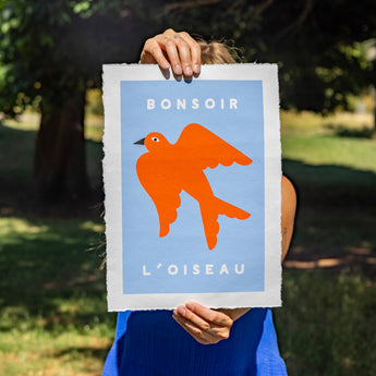 Person holding a framed artwork with an orange bird design against a blue background, outdoors.