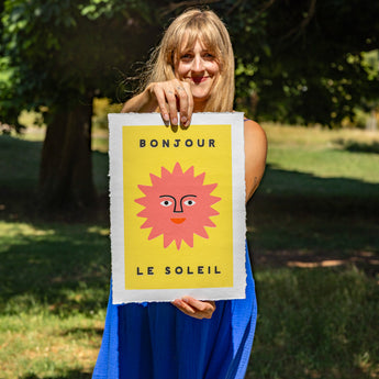 Woman holding a framed artwork with a sun design and 'Bonjour Le Soleil' text in a park.