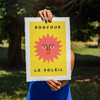 Person holding a poster with a sun illustration and text 'Bonjour Le Soleil' outdoors.